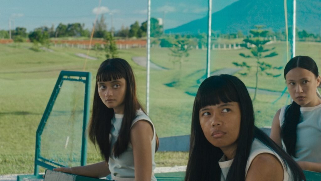 Three young women in matching light-colored shirts sit outdoors against a green landscape with a mountain in the distance.