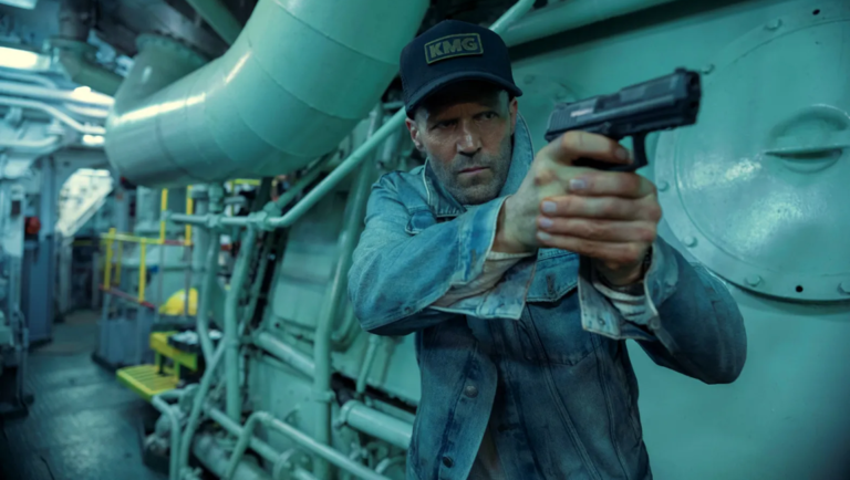 Man in a KMG baseball cap and denim jacket holds a handgun in the engine room of a ship with teal lighting.