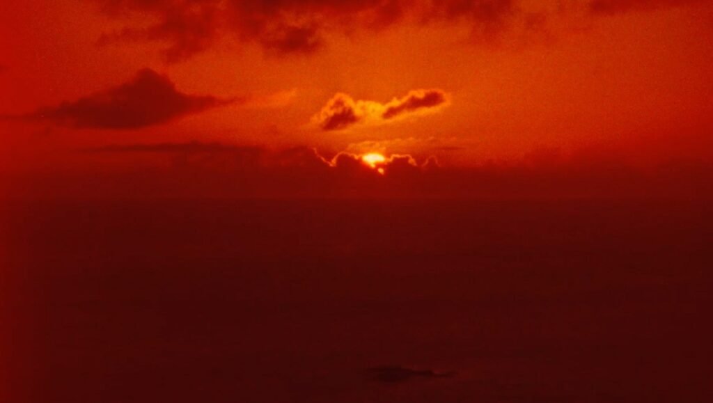 Vibrant red sunset glows behind a thin layer of clouds above a dark, vast horizon.