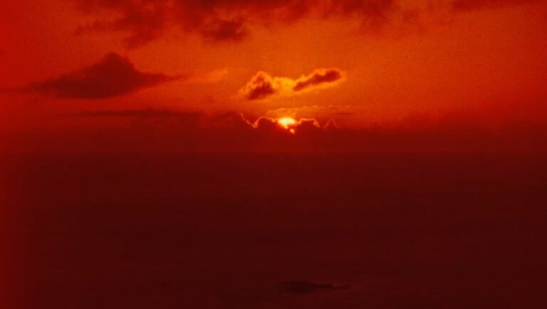 Vibrant red sunset glows behind a thin layer of clouds above a dark, vast horizon.