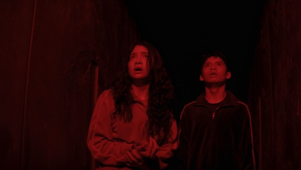 Terrified woman and man look upward in a narrow, dark space bathed in ominous red light.