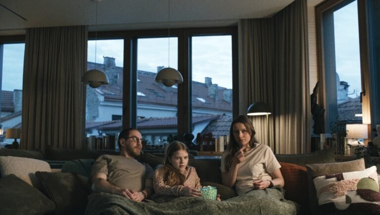A man, a young girl, and a woman sit together on a sofa under a blanket in a dimly lit living room.