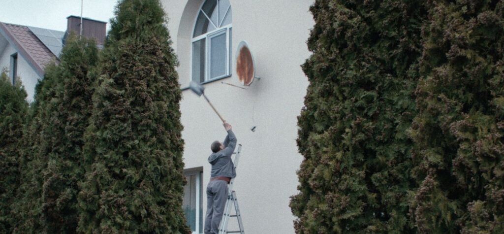 The Way We Are Living: A Conversation with Andrius Blazevicius on How to Divorce During the War Man on a ladder swings a shovel at a rusty satellite dish mounted on the exterior wall of a house between two tall trees.