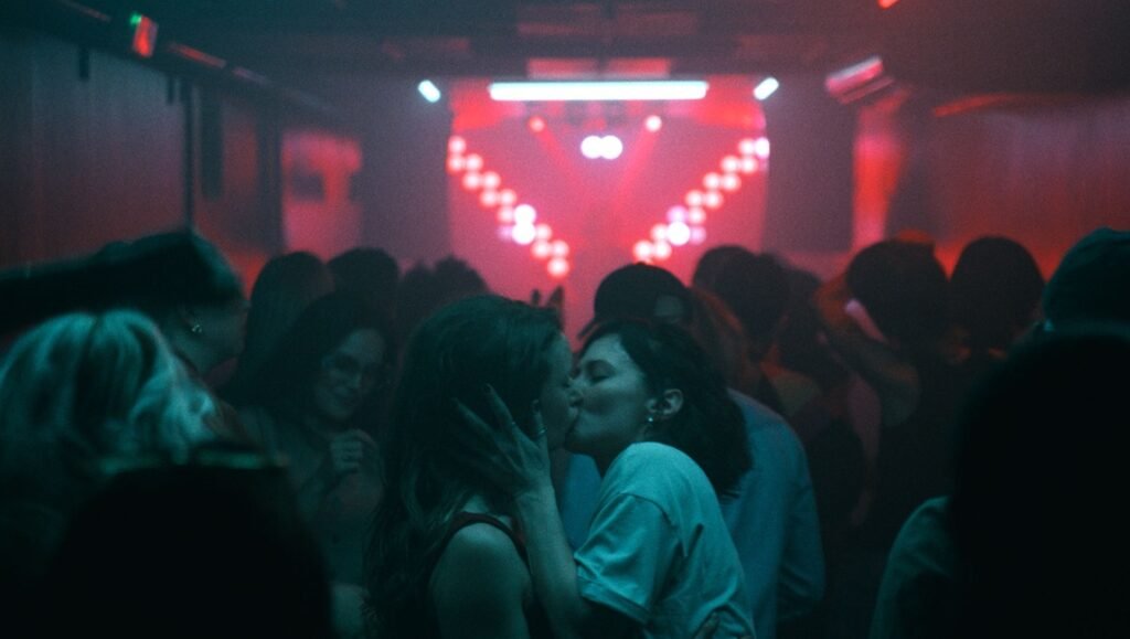 Two women kiss amidst a crowded, dimly lit nightclub featuring glowing red stage lights in the background.