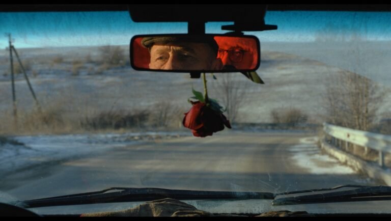 Man wearing a cap reflected in a car rearview mirror above a red rose hanging before a snowy, rural road.