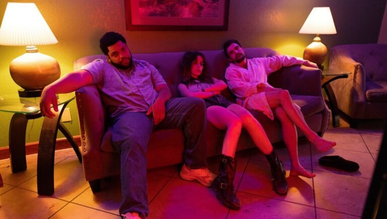 Three people sit on a couch in a dimly lit room with pink and yellow ambient lighting.