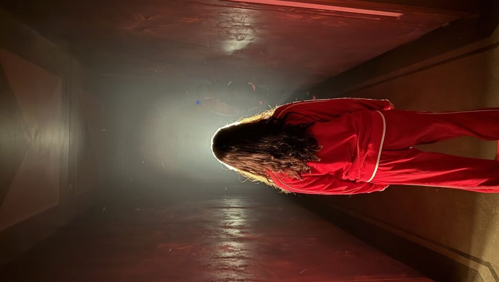 Person with long dark hair wearing a red tracksuit stands in a hazy, dimly lit corridor facing a bright light source.