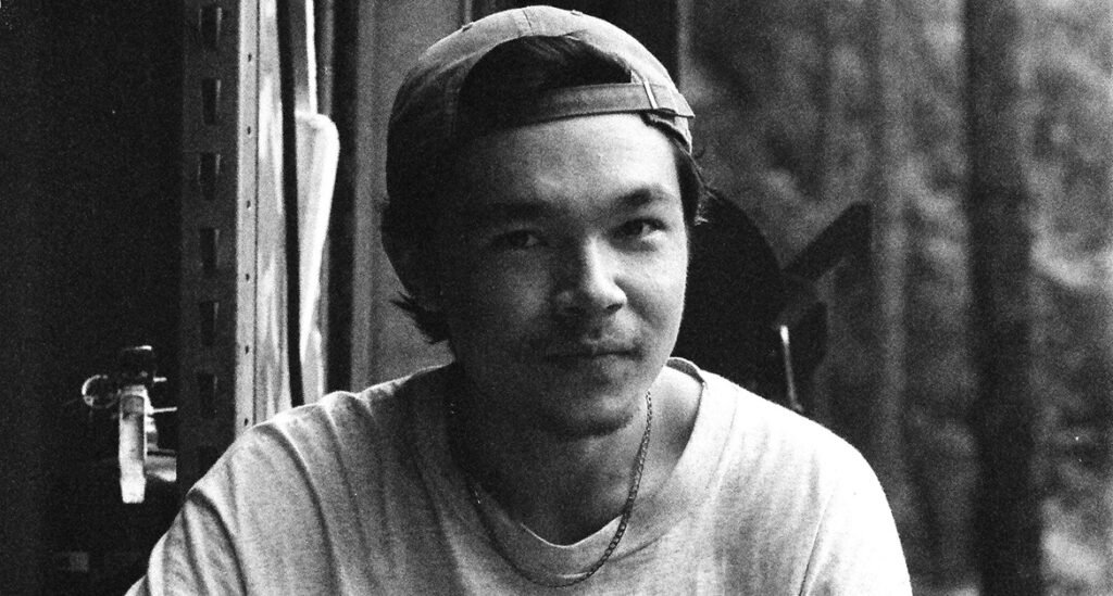 Alex Ullom wearing a baseball cap and a thin chain necklace stares directly at the camera in a grainy black-and-white.