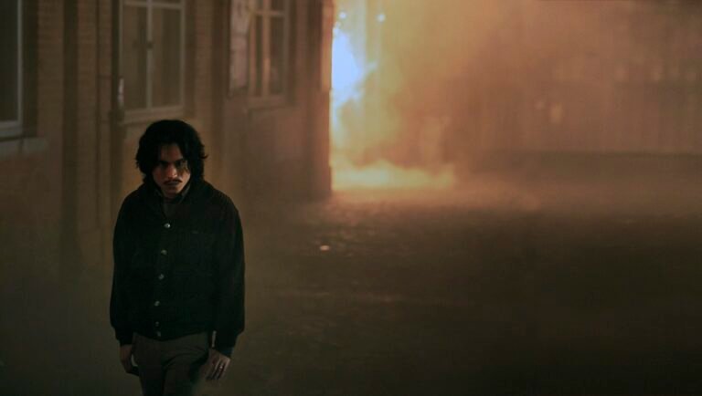 Man with dark hair and a mustache walks through a shadowy alleyway filled with thick, orange-tinted smoke.