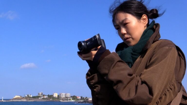 Young woman in a brown jacket filming with a handheld camcorder against a bright blue sky and coastal background.