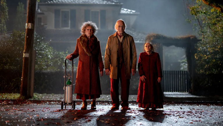 Three elderly people stand on a dark suburban street at night in front of a house.