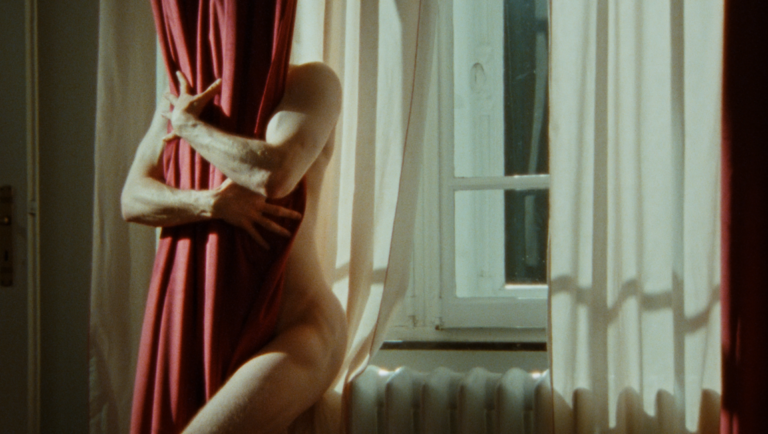 Nude person stands by a window, clutching a heavy deep-red curtain against their torso.