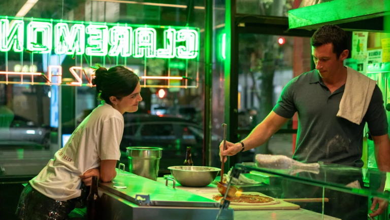 A woman smiles at a man preparing a pizza behind a counter under bright green neon lights reading CLAREMON PIZZA.