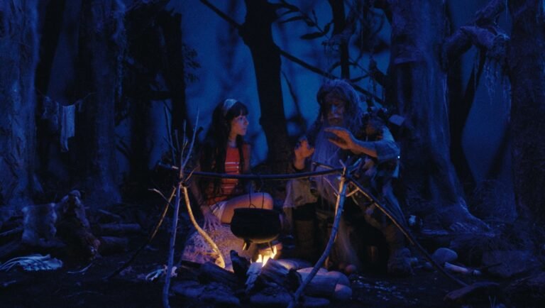 Young girl and a bearded man sit by a campfire in a dark, blue-toned forest.