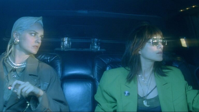 Two women sit in the back of a dark car, one with platinum hair and a tan coat, the other wearing a green blazer and shades.