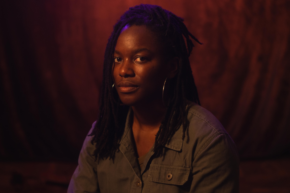 Shatara Michelle Ford with dreadlocks and hoop earrings against a warm, dimly lit red background.