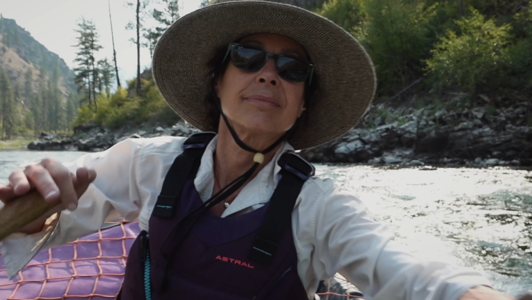 Close-up of a smiling rower in a wide-brimmed hat and Astral life vest navigating a river past rocky, forested banks.