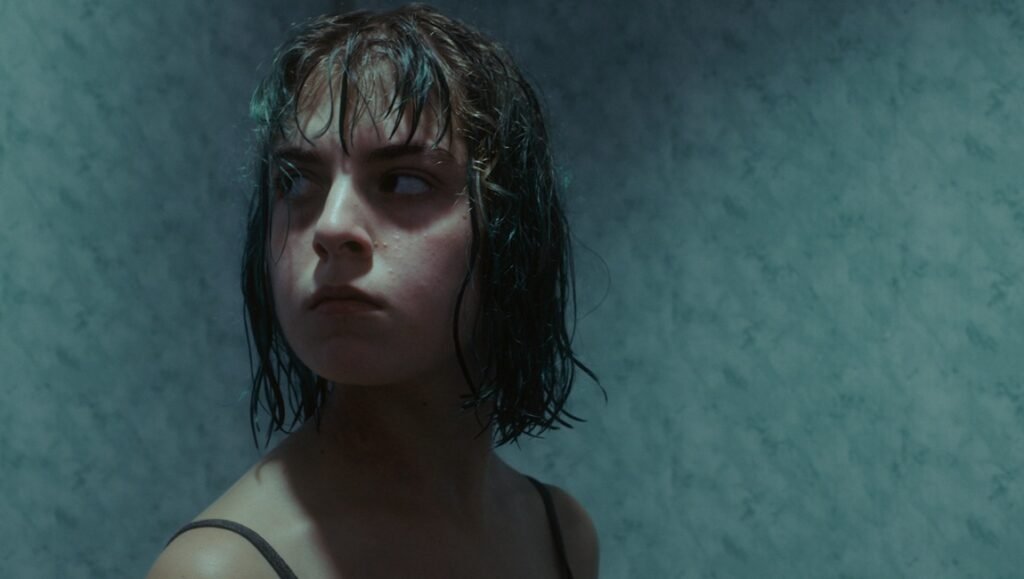 Young person with wet, dark hair stares intensely toward the left side against a muted teal wall.