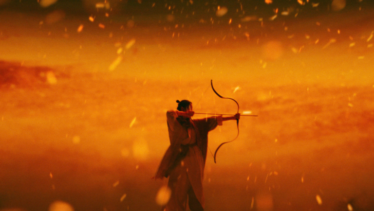 Archer in traditional robes aims a bow amidst golden embers against a hazy, warm-toned background.