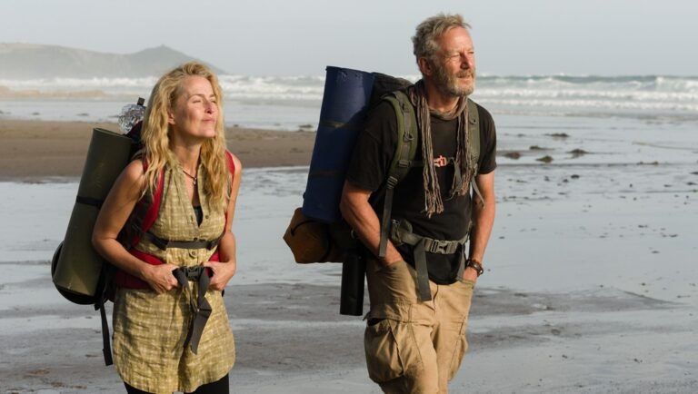 A woman and a man with large hiking backpacks walk along a sandy beach near the ocean.