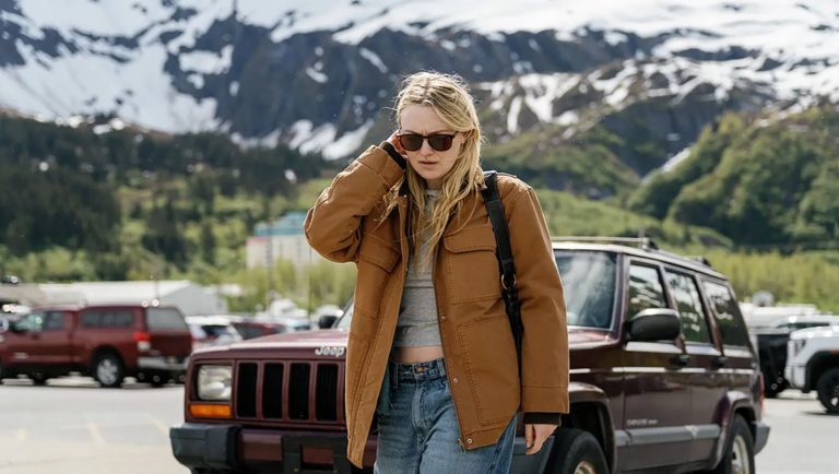 Blonde woman wearing sunglasses and a brown jacket walks past a maroon Jeep in front of snow-capped mountains.