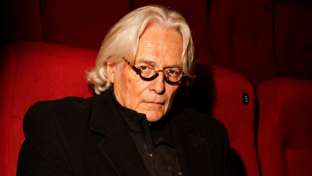 Man with silver hair and glasses sitting in a red cinema seat.