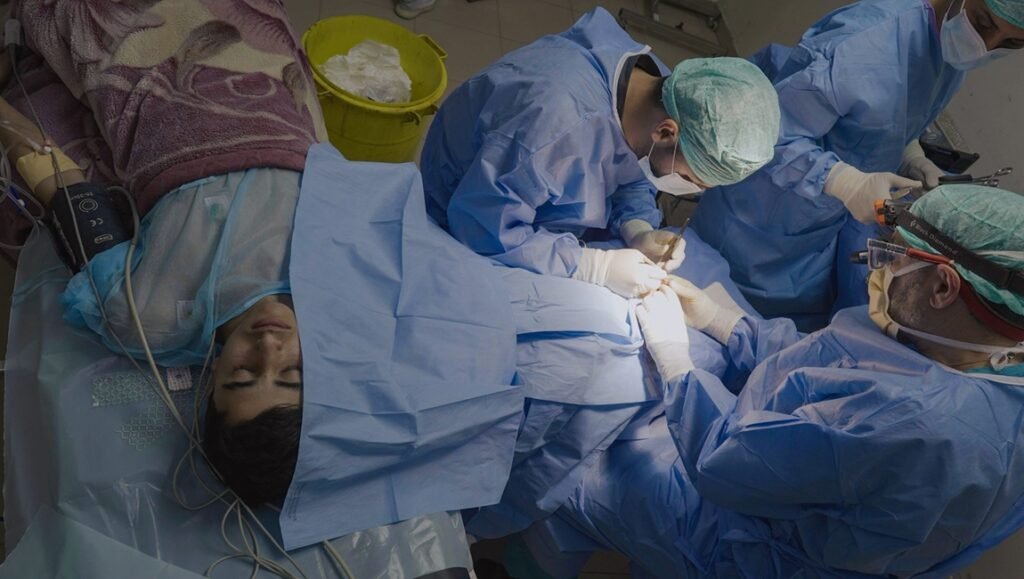 Patient lies on an operating table while surgeons in blue gowns and protective gear work together during surgery.