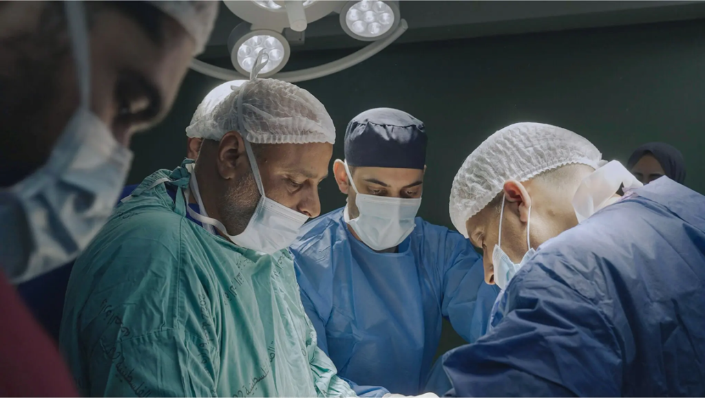 Medical team in surgical scrubs and caps concentrates intently while performing a procedure under bright lights.