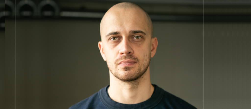 Andrius Blazevicius with a shaved head and light stubble stares directly at the camera against a dark background.