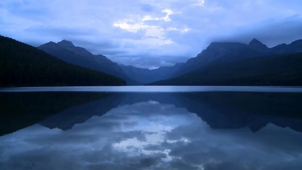 Dark mountains reflect perfectly on the calm, blue-tinted surface of Bowman Lake under a cloudy twilight sky.