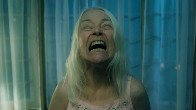 Woman with pale hair and blank white eyes screams in a dimly lit room with blue curtains.