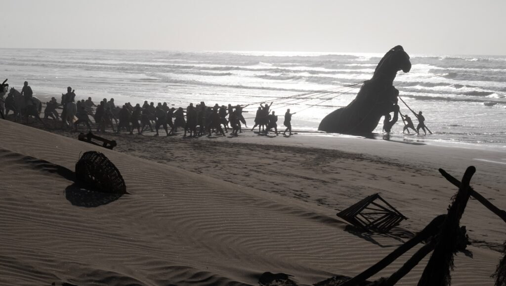 Large group of silhouetted figures pulls ropes to drag a massive wooden horse sculpture across a sunlit beach.