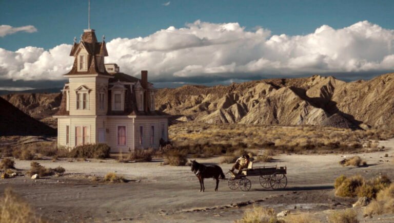 A person drives a horse-drawn carriage past a Victorian-style house in a dry, mountainous desert landscape.