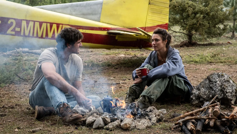 A man and woman sit by a campfire in front of a yellow and red airplane tail in a wooded area.