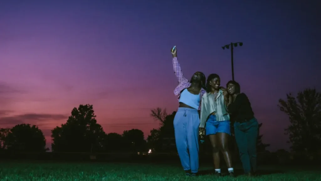 Three women laughing and posing for a selfie in a grassy field against a vibrant purple and orange twilight sky.