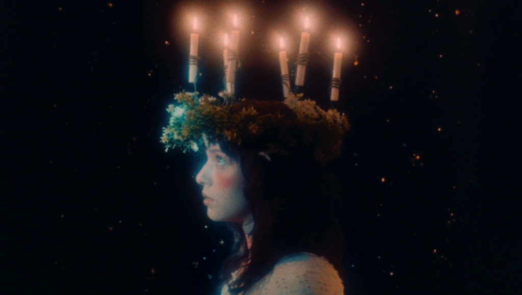 Profile of a girl wearing a dark floral crown topped with five glowing candles against a starry night background.