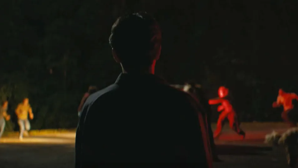 Person viewed from behind watches figures run through a dark landscape illuminated by glowing red and yellow neon lights.