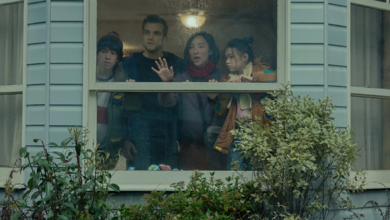 Four people look out through a rain-streaked window from inside a house with light blue siding.