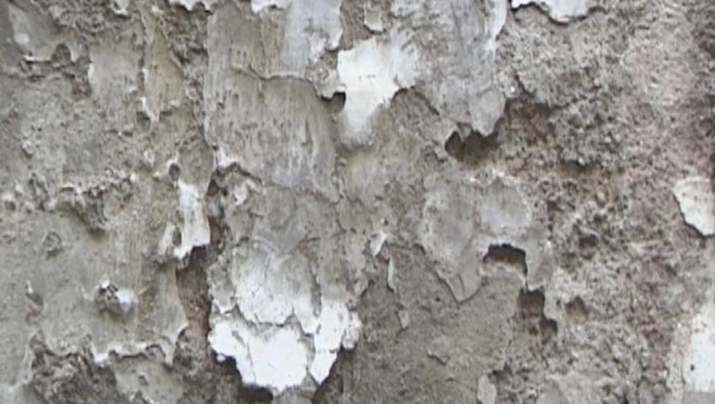 Weathered concrete wall featuring patches of peeling white paint over a textured gray surface.