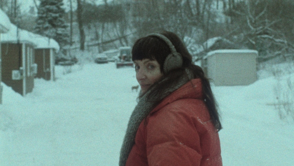 Woman with dark bangs and earmuffs wearing a red puffer jacket looks over her shoulder in a snowy residential setting.