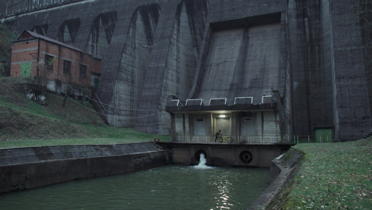 A person with a bicycle stands on a concrete platform before a massive dam structure with water flowing from a pipe.