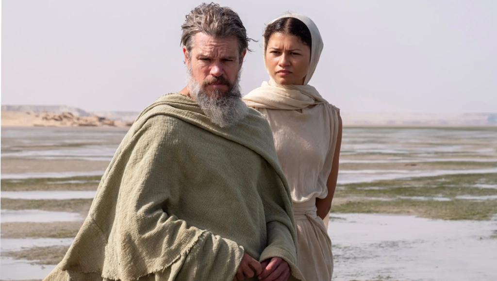 Bearded man in a sage cloak and woman in a white headscarf stand on a desolate, light-colored landscape.