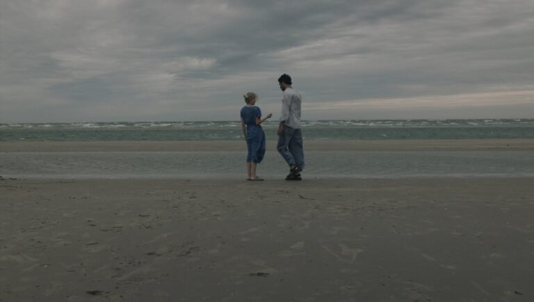 Young couple stands on a wet sandy beach facing a turbulent sea under a heavy, overcast September sky.