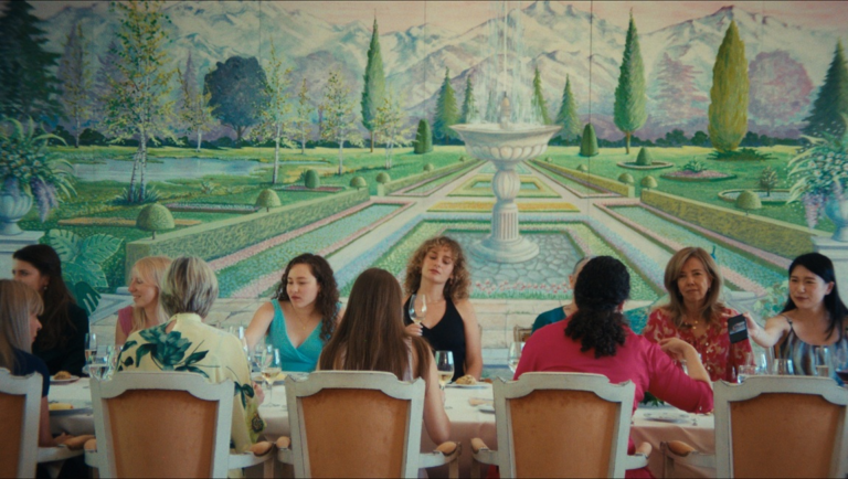 Diverse group of women sitting at a formal dining table in front of a painted landscape mural of a garden and fountain.