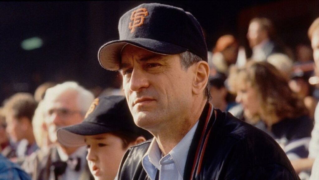 Robert De Niro wears a San Francisco Giants baseball cap while sitting in a stadium crowd next to a young boy.
