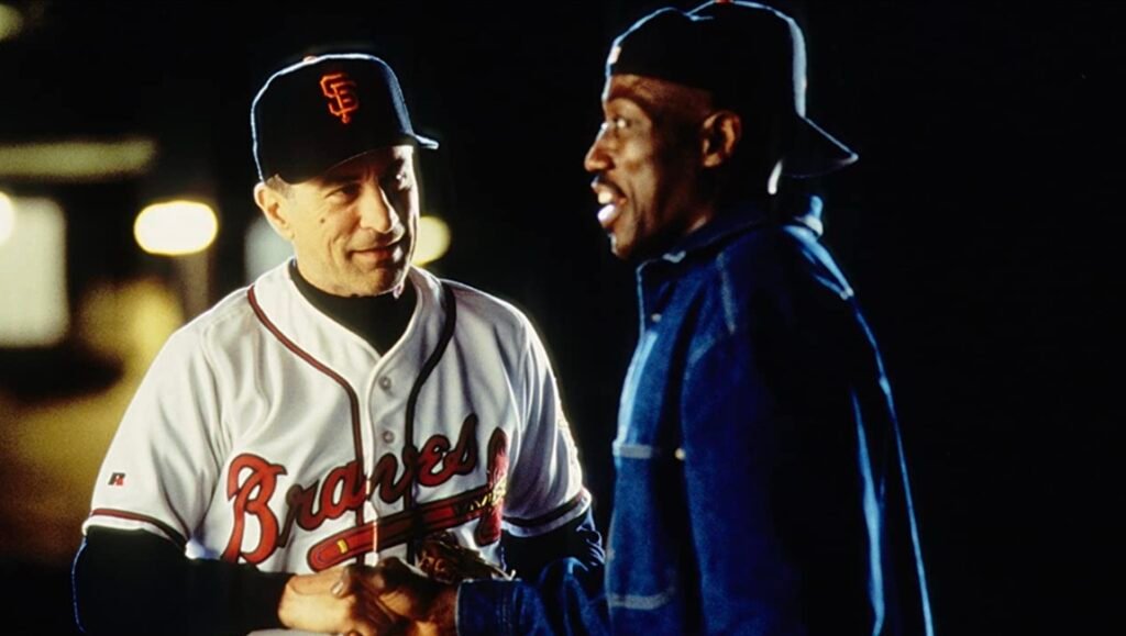Robert De Niro in a Braves baseball uniform smiles while shaking hands with Wesley Snipes at night.