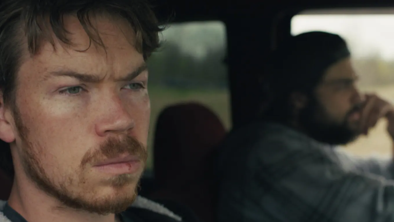 Will Poulter sits in the passenger seat of a car with a serious expression while another man drives.