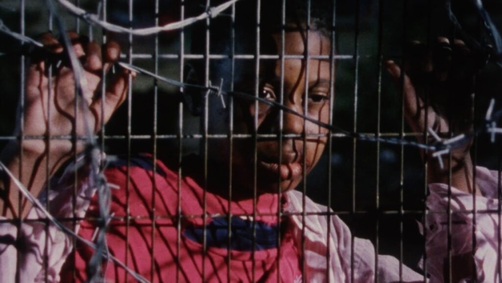 There’s No Solution, But It Has to Come: An Interview with Ngozi Onwurah on Welcome II the Terrordome Young person looking through a metal wire fence with barbed wire.