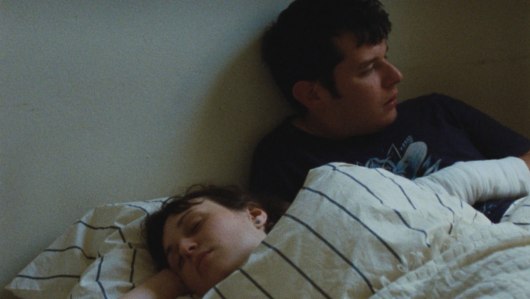 A man looking away while a woman sleeps beside him under a striped blanket in a dimly lit room.