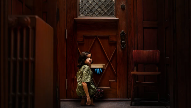 A young child crouches before a dark wooden door, looking back as a mysterious blue hand reaches through the mail slot.